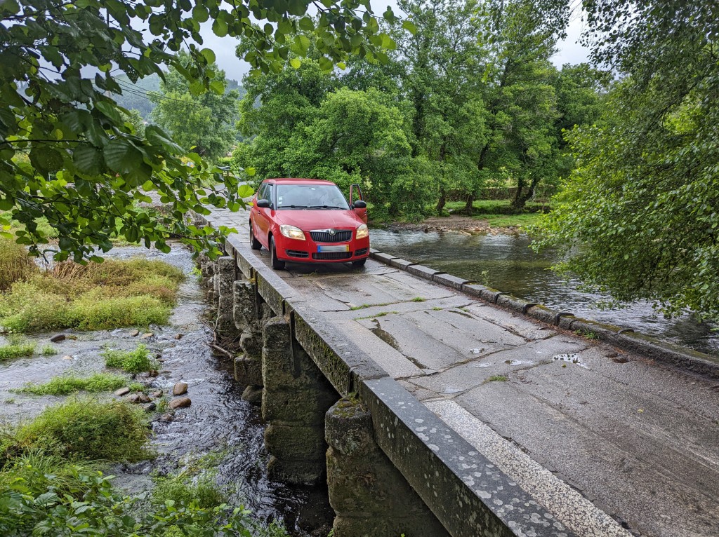 Ein rotes Auto fährt über eine steinerne Brücke über einen reißenden Fluss.