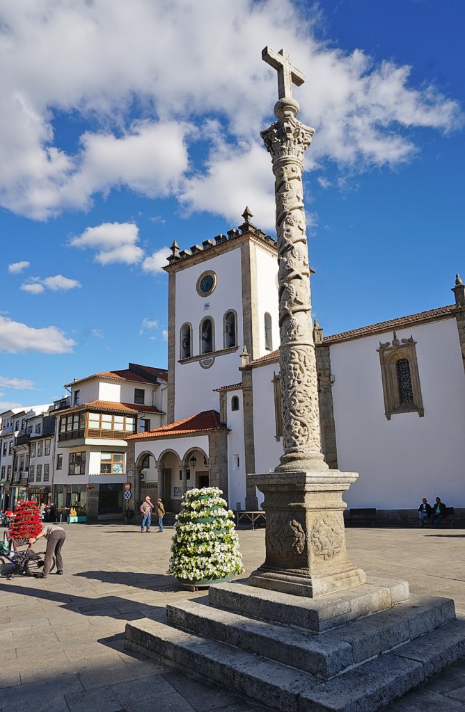 Ein Säule mit Skulpturen steht auf einem Platz vor einer Kirche im Sonnenschein