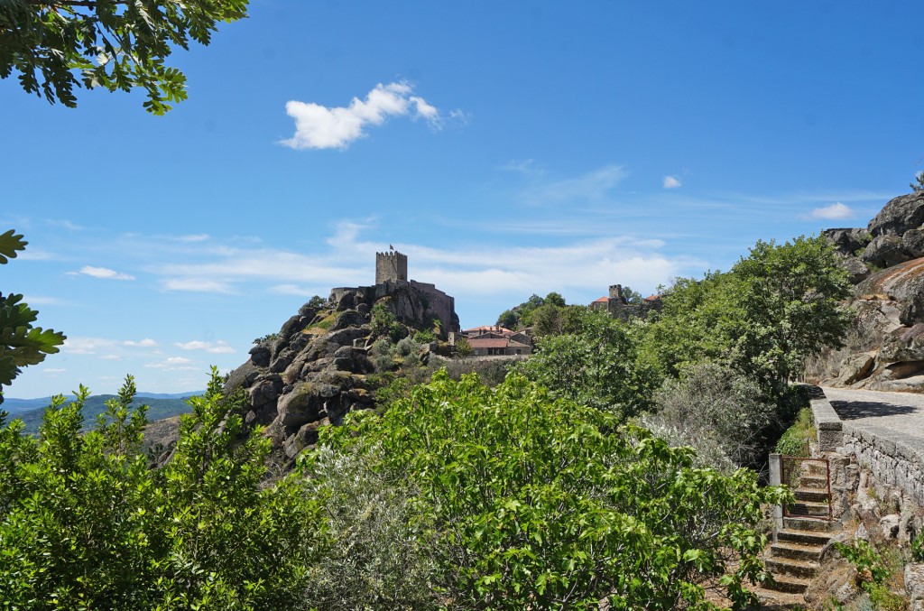 Eine Burg thront auf einem Felsen, umgeben von grünen Bäumen und einem blauen Himmel