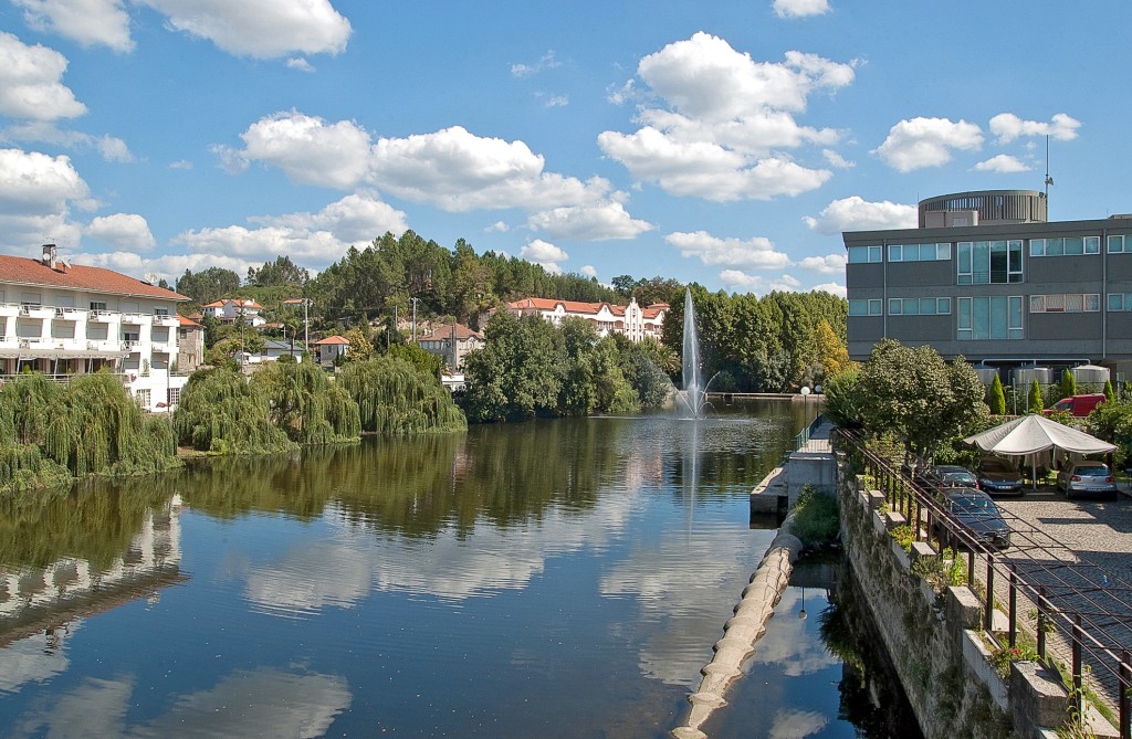 Ein See mit Bäumen und Gebäuden spiegelt sich im Wasser unter einem blauen Himmel.