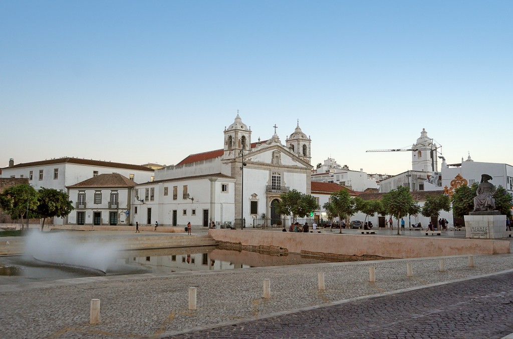 White buildings and a church tower are visible in a town square with a fountain
