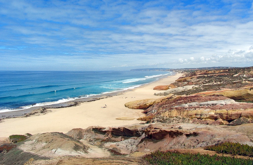 Weite Sandküste mit rotem Fels und blauem Meer unter blauem Himmel