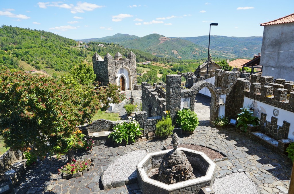 Stone fountain sits in a garden with views of rolling green hills and a church.