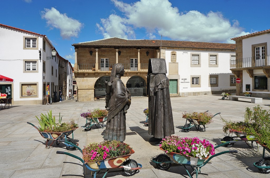 Bronze statues stand in a square surrounded by buildings and colorful wheelbarrows