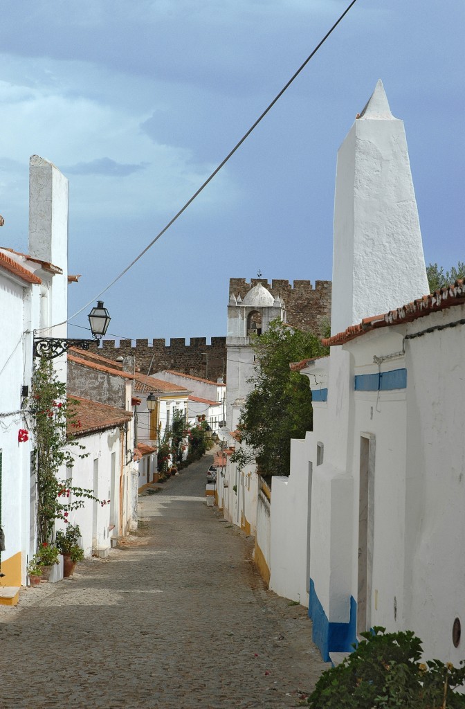 Eine malerische gepflasterte Gasse mit weißen Häusern und einer Burg im Hintergrund.