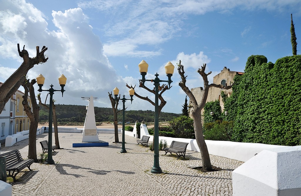 Ein Platz mit Bäumen Bänken und einem Denkmal unter blauem Himmel.