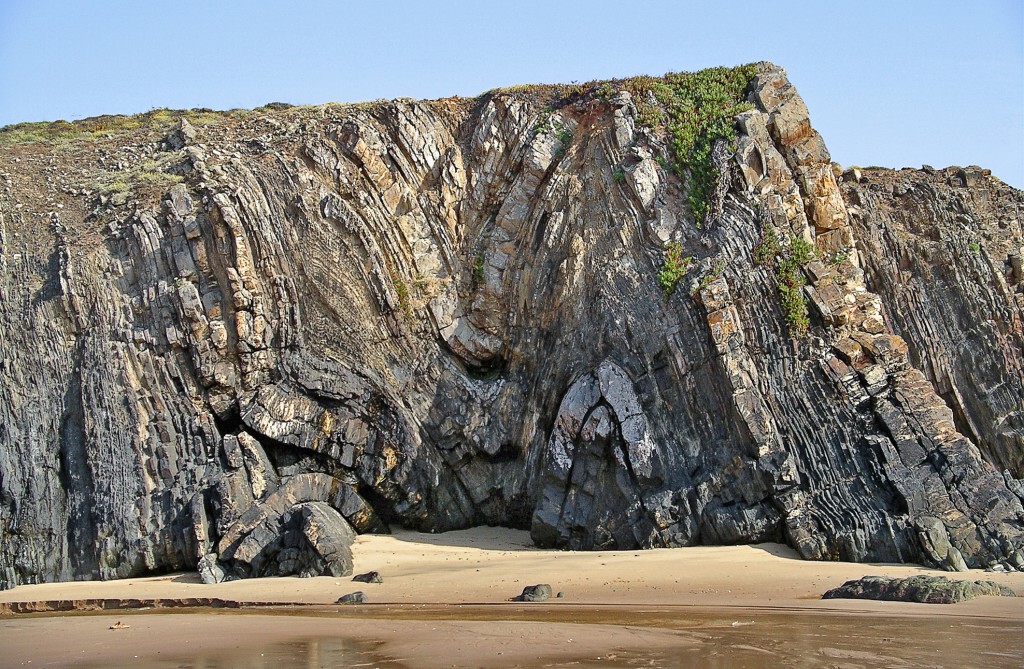 Felswand mit dunklen Schichten und Strand darunter.