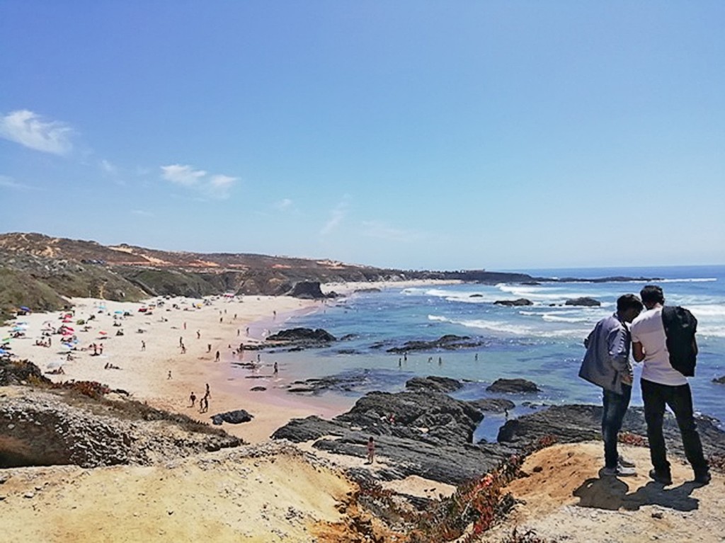 Zwei Personen betrachten einen sonnigen Strand mit Wellen und Felsen im Hintergrund