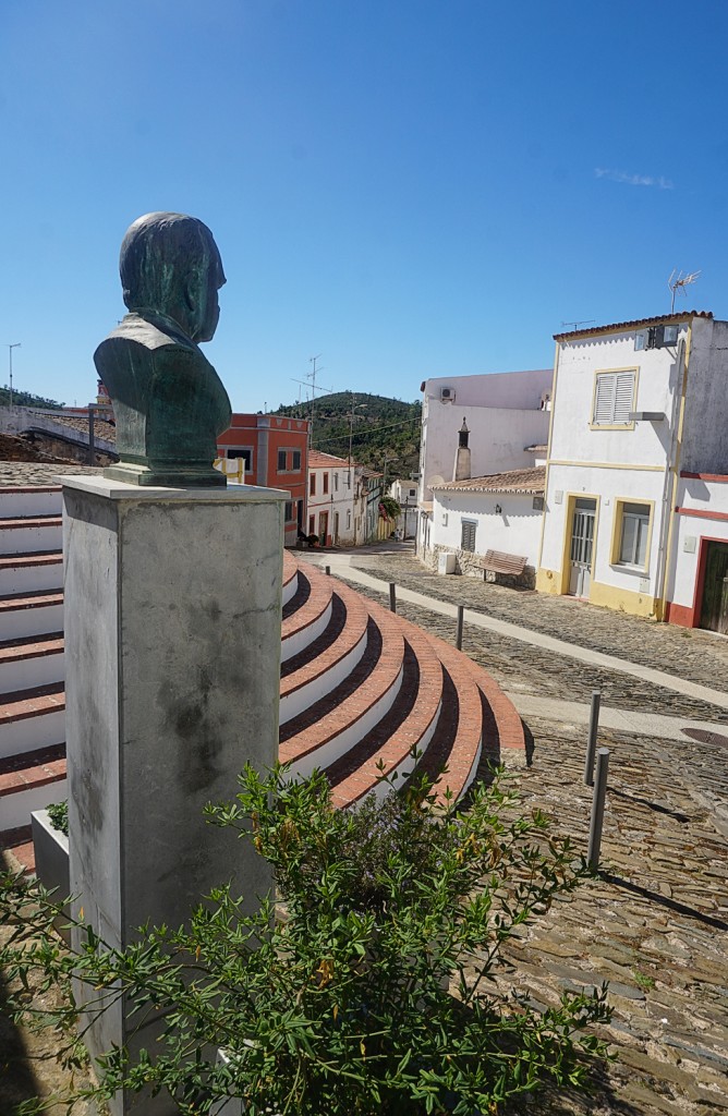 Eine Bronzebüste blickt über eine Treppe in eine malerische Stadtlandschaft.